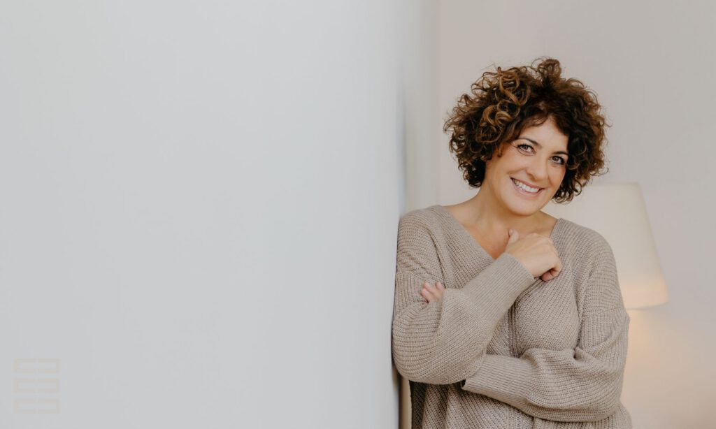Familien- und Wirtschaftsmediatorin Jasmin Hirnich steht freundlich lachend an einer Wand. Im Hintergrund eine Stehleuchte.