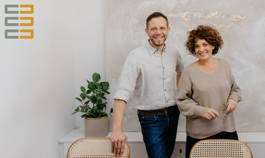 Mediatoren Sören Peter und Jasmin Hirnich stehen zusammen vor zwei Stühlen. Im Hintergrund eine Pflanze und ein Bild. Links befindet sich das Logo.