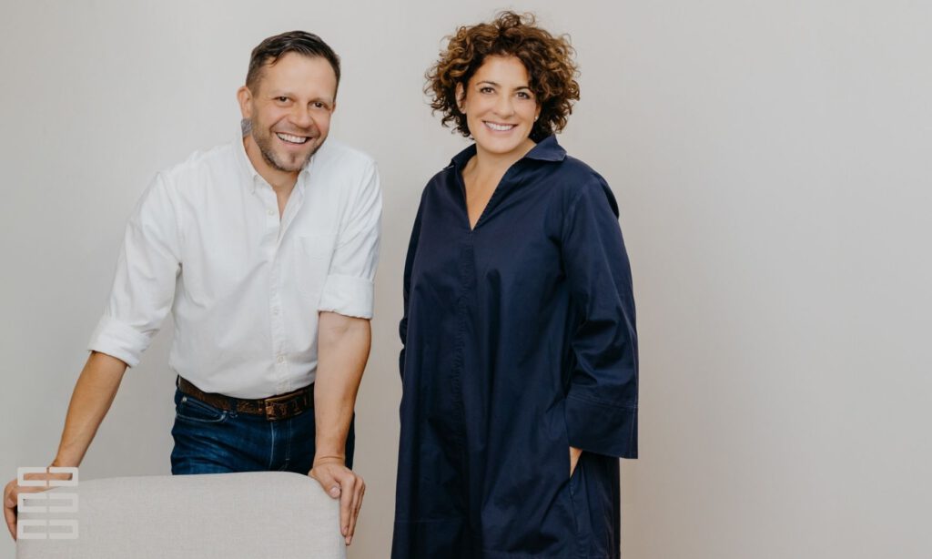 Wirtschaftsmediatoren Sören Peter und Jasmin Hirnich stehen lässig, lachend vor einer neutralen Wand.